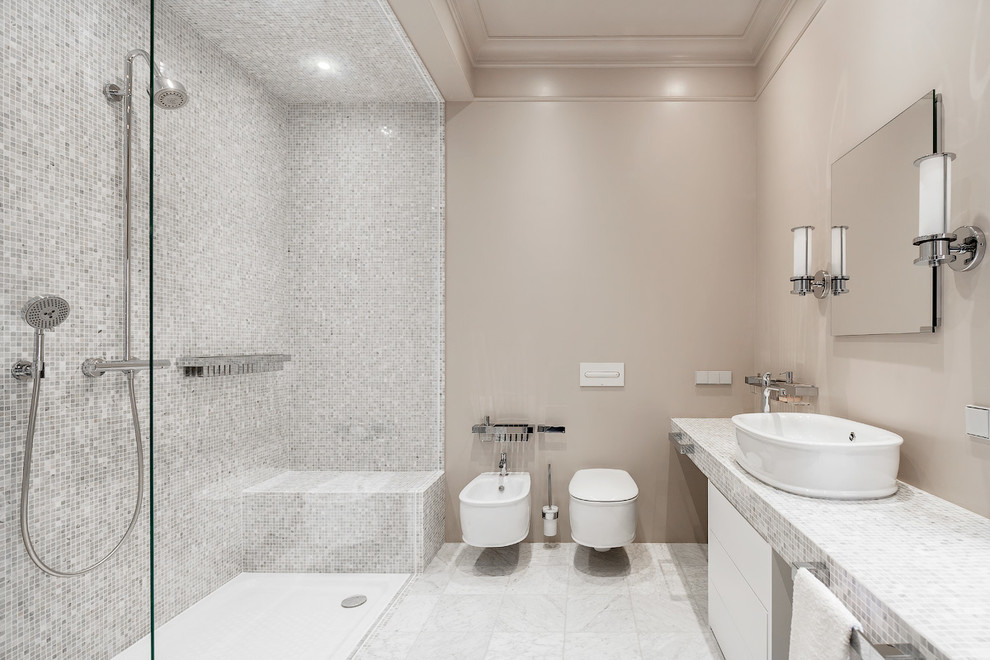 Cette photo montre une salle de bain principale chic avec un placard à porte plane, des portes de placard blanches, une douche à l'italienne, WC suspendus, un carrelage blanc, un carrelage de pierre, un mur beige, un sol en marbre, une vasque et un plan de toilette en carrelage.