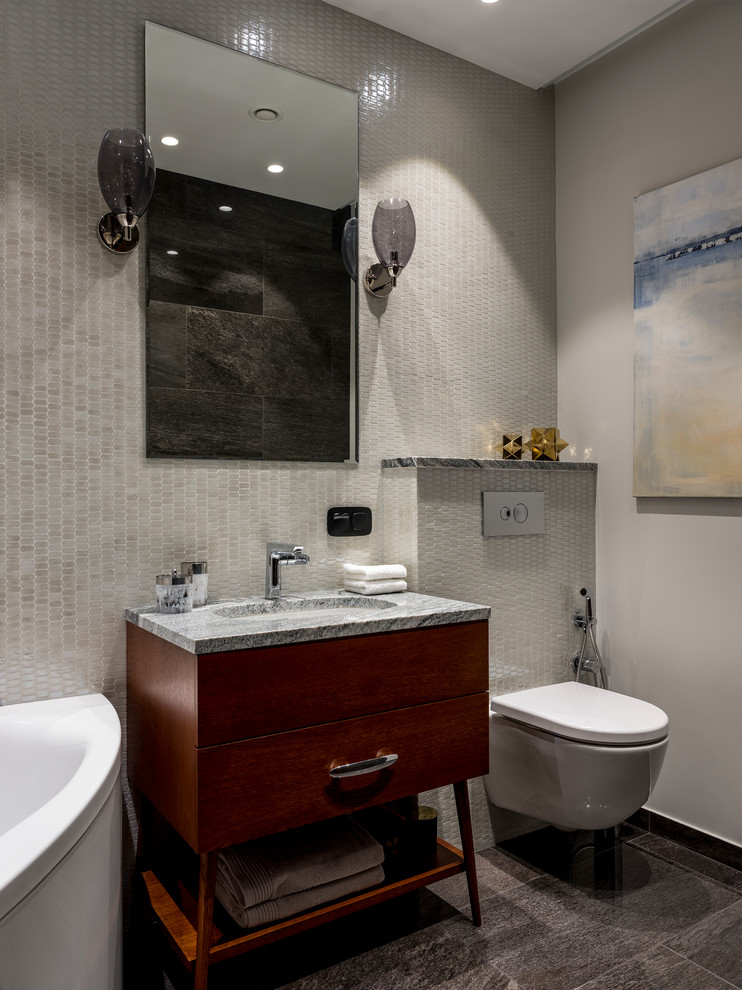 Modelo de cuarto de baño principal contemporáneo con armarios con paneles lisos, puertas de armario de madera en tonos medios, bañera esquinera, sanitario de pared, baldosas y/o azulejos grises, lavabo bajoencimera, encimera de granito, suelo gris y encimeras grises