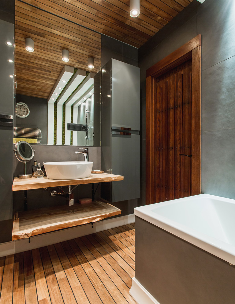 Modelo de cuarto de baño principal actual con armarios abiertos, combinación de ducha y bañera, baldosas y/o azulejos grises, suelo de madera pintada, lavabo sobreencimera, suelo marrón y encimera de madera