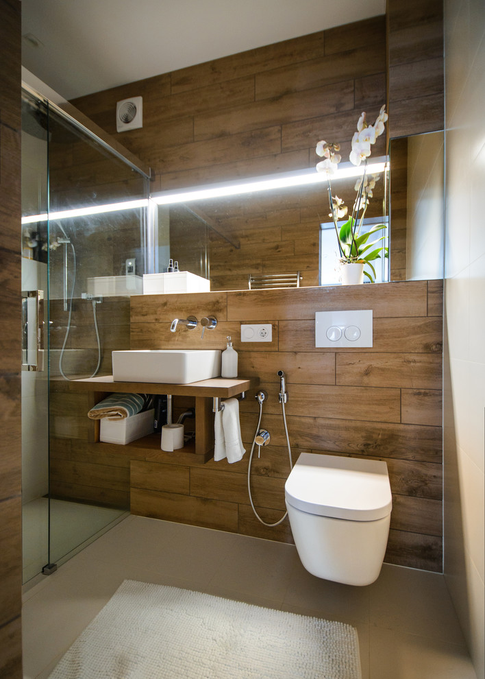 Photo of a small contemporary shower room bathroom in Novosibirsk with open cabinets, medium wood cabinets, an alcove bath, grey tiles, white tiles, brown tiles, porcelain flooring, a vessel sink, wooden worktops, a wall mounted toilet, porcelain tiles, brown worktops and feature lighting.