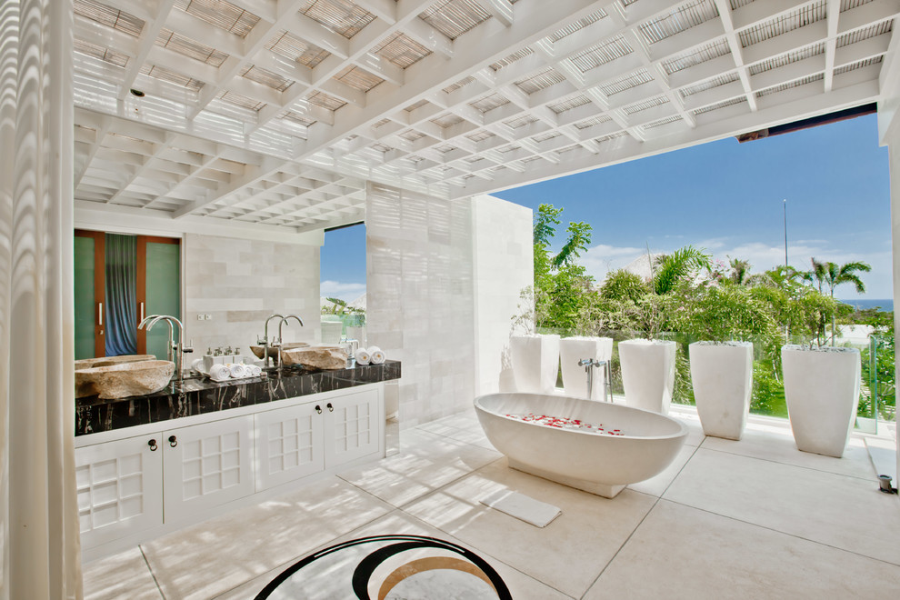 На фото: главная ванная комната в морском стиле с фасадами с утопленной филенкой, белыми фасадами, отдельно стоящей ванной, белыми стенами, черной столешницей, серой плиткой, настольной раковиной и бежевым полом