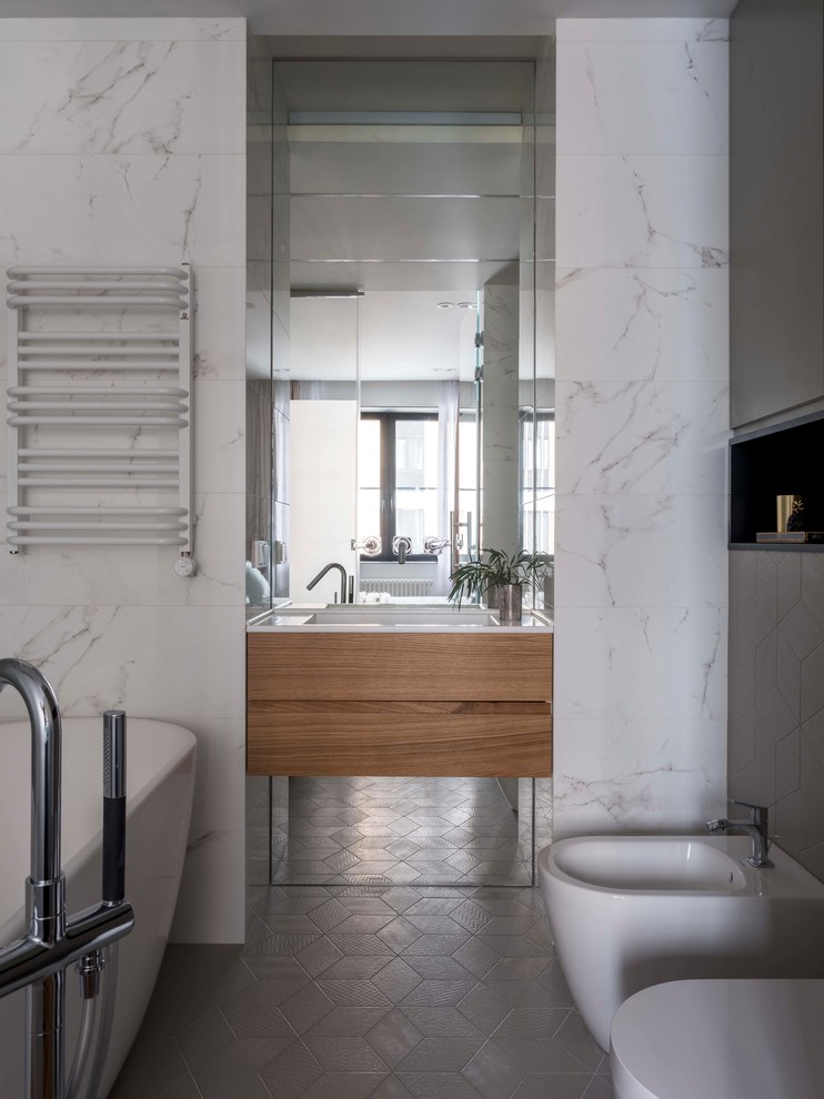 Идея дизайна: главная ванная комната в современном стиле с отдельно стоящей ванной, плоскими фасадами, фасадами цвета дерева среднего тона, биде, белой плиткой, керамогранитной плиткой, серым полом, белой столешницей, серыми стенами и монолитной раковиной