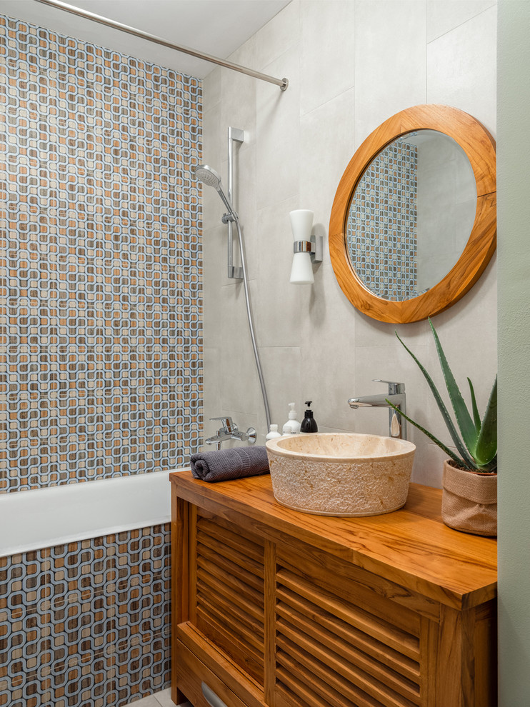Cette image montre une salle d'eau design en bois brun avec un placard à porte persienne, un carrelage gris, un carrelage multicolore, un mur gris, une vasque, un plan de toilette en bois, un plan de toilette marron, une baignoire en alcôve et un combiné douche/baignoire.