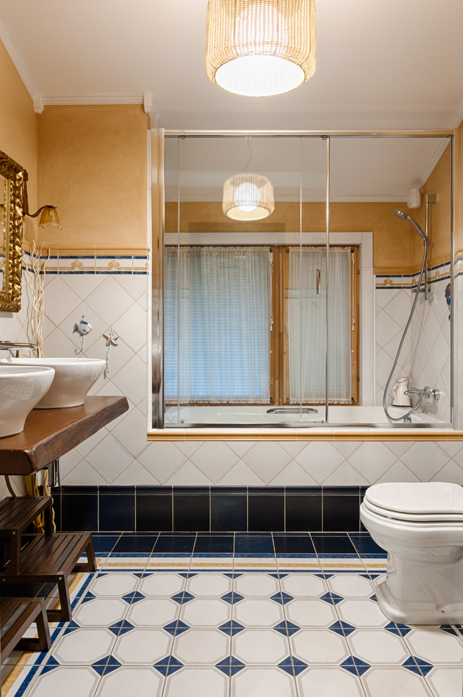 Exempel på ett rustikt brun brunt en-suite badrum, med ett badkar i en alkov, en dusch/badkar-kombination, en toalettstol med separat cisternkåpa, vit kakel, blå kakel, gula väggar, ett fristående handfat, flerfärgat golv, träbänkskiva och dusch med skjutdörr