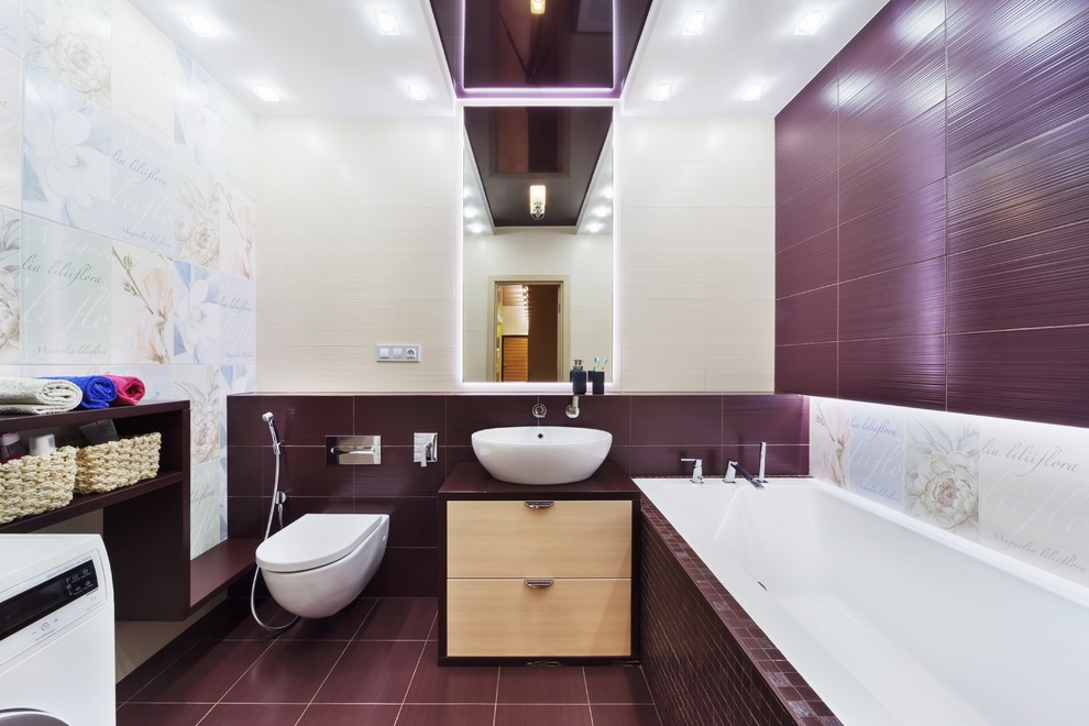 Modelo de cuarto de baño contemporáneo con armarios con paneles lisos, puertas de armario beige, bañera encastrada, sanitario de pared, baldosas y/o azulejos multicolor, paredes púrpuras, aseo y ducha, lavabo sobreencimera y suelo violeta