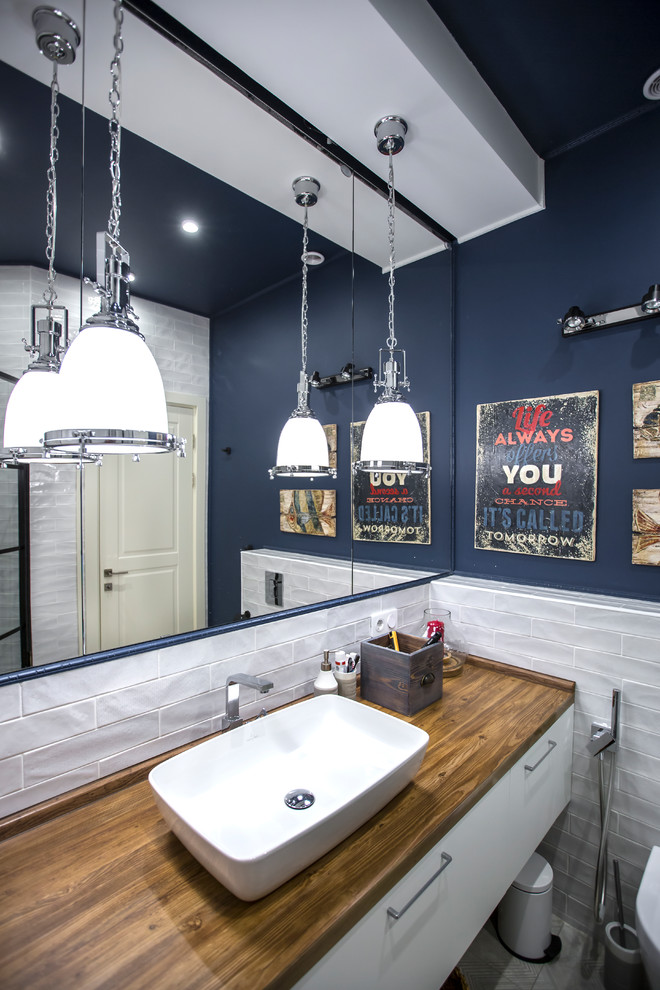 Bild på ett mellanstort funkis brun brunt badrum med dusch, med en dusch i en alkov, en vägghängd toalettstol, vit kakel, keramikplattor, blå väggar, klinkergolv i keramik, grått golv, dusch med gångjärnsdörr, släta luckor, vita skåp, ett fristående handfat och träbänkskiva