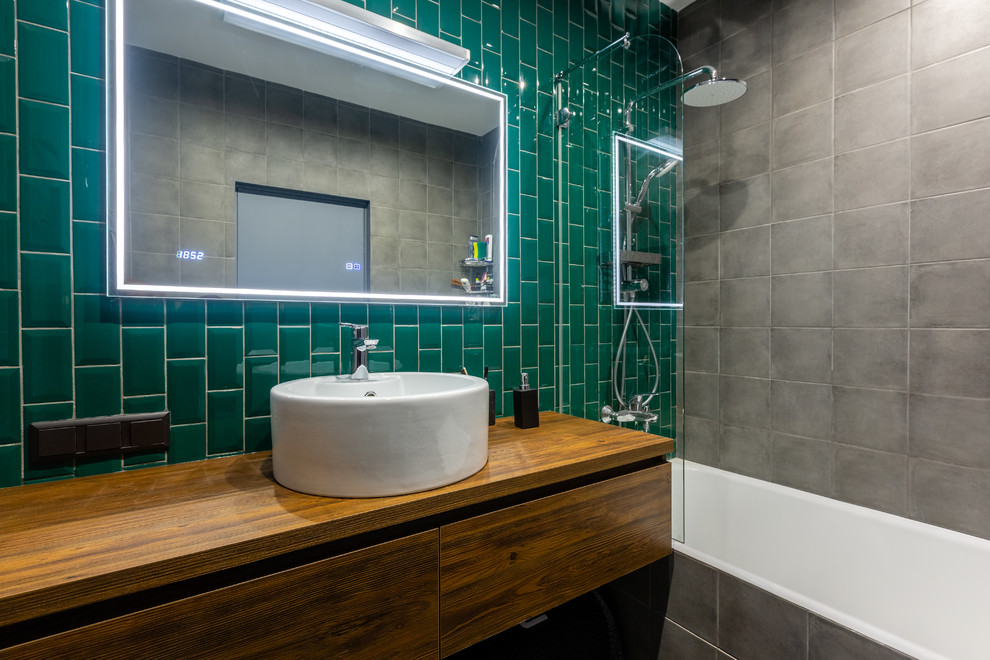 Diseño de cuarto de baño urbano con armarios con paneles lisos, puertas de armario de madera oscura, bañera empotrada, combinación de ducha y bañera, baldosas y/o azulejos verdes, lavabo sobreencimera, encimera de madera y espejo con luz
