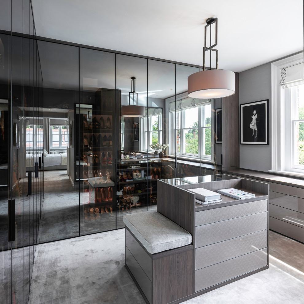 Photo of a large contemporary gender neutral dressing room in Hertfordshire with glass-front cabinets, dark wood cabinets, carpet and grey floors.