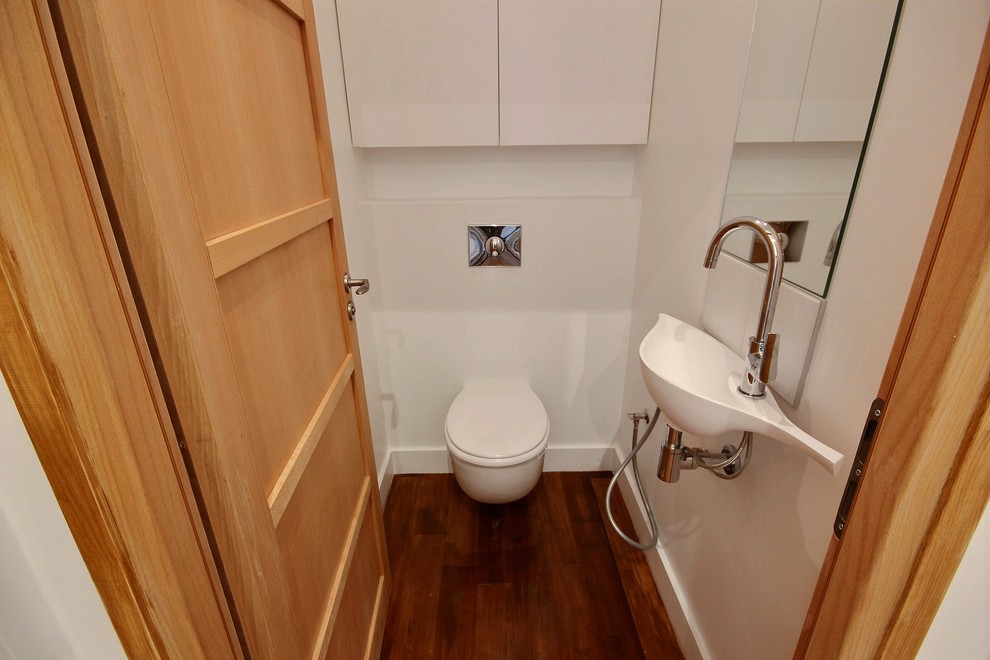 Aménagement d'un petit WC suspendu industriel avec des portes de placard blanches, un mur blanc, un sol en bois brun et un lavabo suspendu.