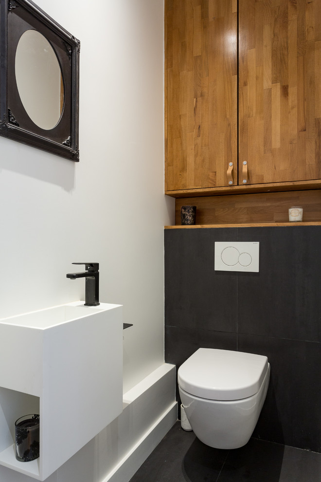 Exemple d'un petit WC suspendu tendance en bois clair avec un placard à porte plane, un carrelage noir, des carreaux de céramique, un mur blanc, un sol en carrelage de céramique, un lavabo suspendu, un plan de toilette en surface solide, un sol noir et un plan de toilette blanc.