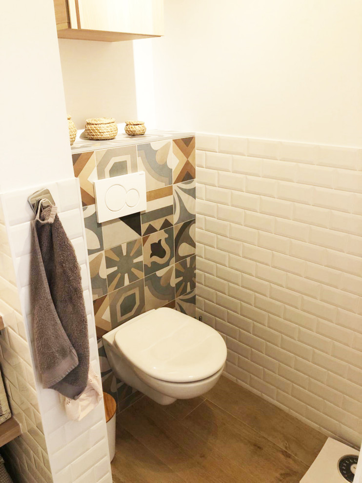 Bild på ett litet lantligt toalett, med bruna skåp, en vägghängd toalettstol, flerfärgad kakel, cementkakel, vita väggar och brunt golv