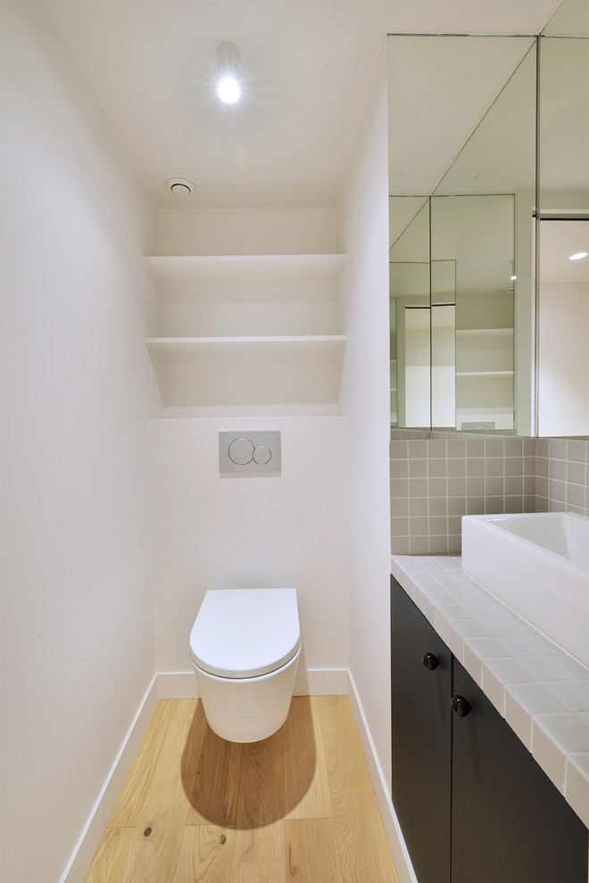 Bild på ett mellanstort funkis grå grått toalett, med luckor med profilerade fronter, svarta skåp, en vägghängd toalettstol, grå kakel, keramikplattor, vita väggar, ljust trägolv, ett nedsänkt handfat och kaklad bänkskiva