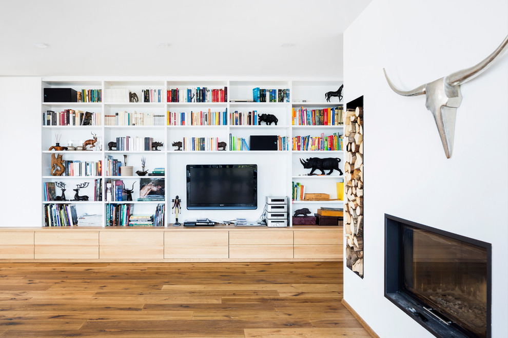 Modelo de biblioteca en casa abierta escandinava grande con paredes blancas, suelo de madera oscura, estufa de leña, marco de chimenea de yeso, televisor colgado en la pared y suelo marrón