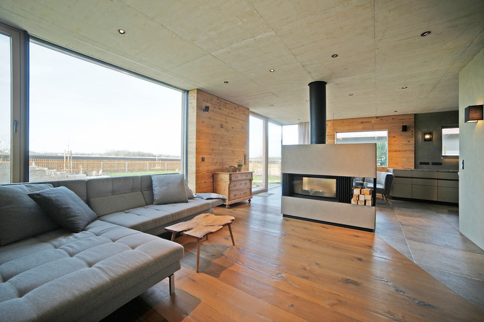 Cette photo montre un grand salon tendance ouvert avec un sol en bois brun, une cheminée double-face, un manteau de cheminée en plâtre, un téléviseur fixé au mur, une salle de réception, un mur gris et un sol marron.