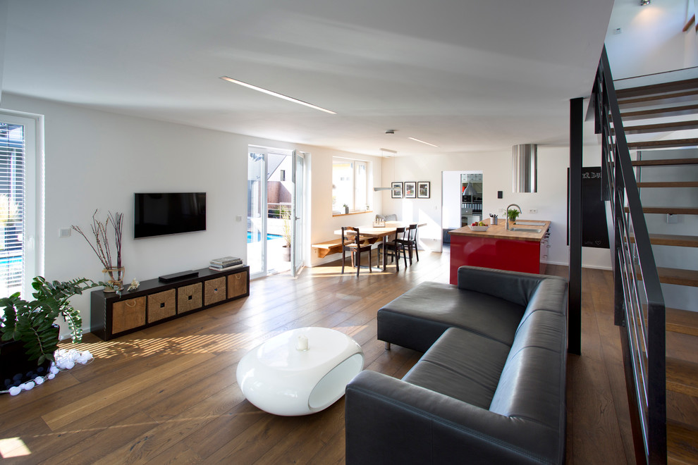 Diseño de salón abierto grande con paredes blancas, suelo de madera en tonos medios y televisor colgado en la pared