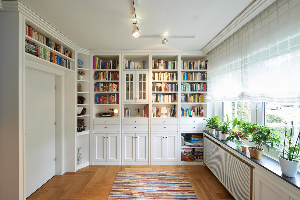 Modelo de sala de estar con biblioteca cerrada tradicional renovada con paredes blancas, suelo de madera en tonos medios y suelo marrón