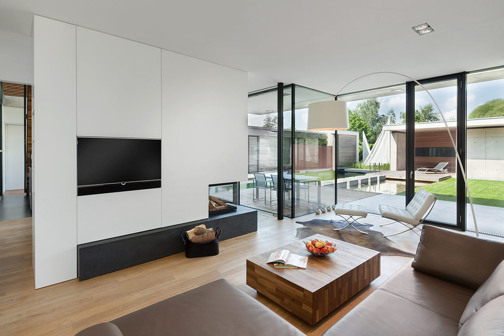 Immagine di un soggiorno design aperto con pareti bianche, pavimento in legno massello medio, camino bifacciale e parete attrezzata