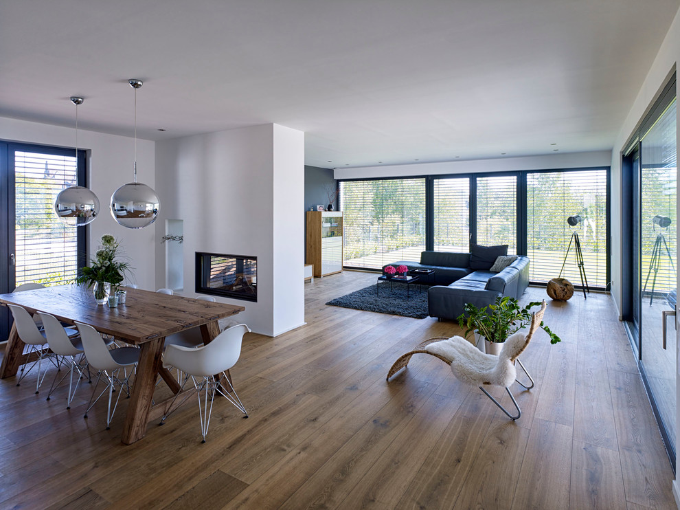 Diseño de salón abierto moderno grande con paredes blancas, suelo de madera en tonos medios, chimenea de doble cara y marco de chimenea de yeso