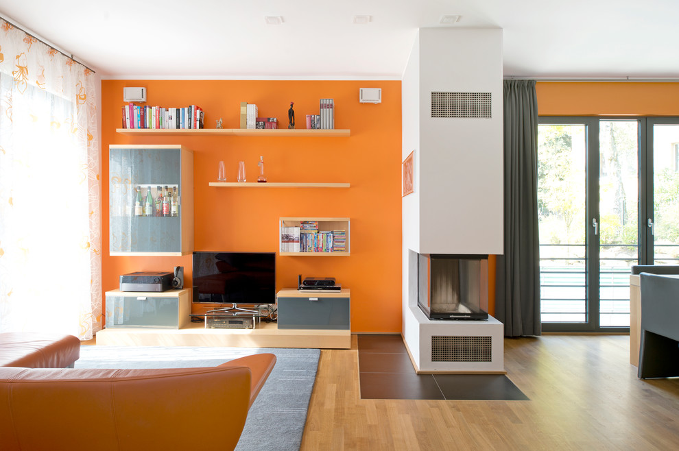 Immagine di un soggiorno minimal di medie dimensioni e aperto con pareti arancioni, parquet chiaro, camino bifacciale, cornice del camino in cemento e parete attrezzata