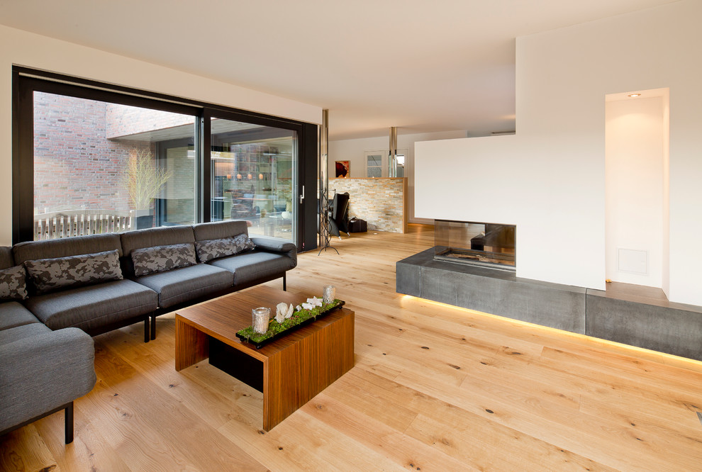 Ejemplo de sala de estar actual con paredes blancas, suelo de madera clara, chimenea de esquina y marco de chimenea de hormigón