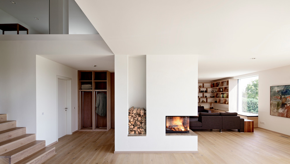 Esempio di un grande soggiorno minimalista aperto con pareti bianche, parquet chiaro, camino bifacciale e cornice del camino in intonaco