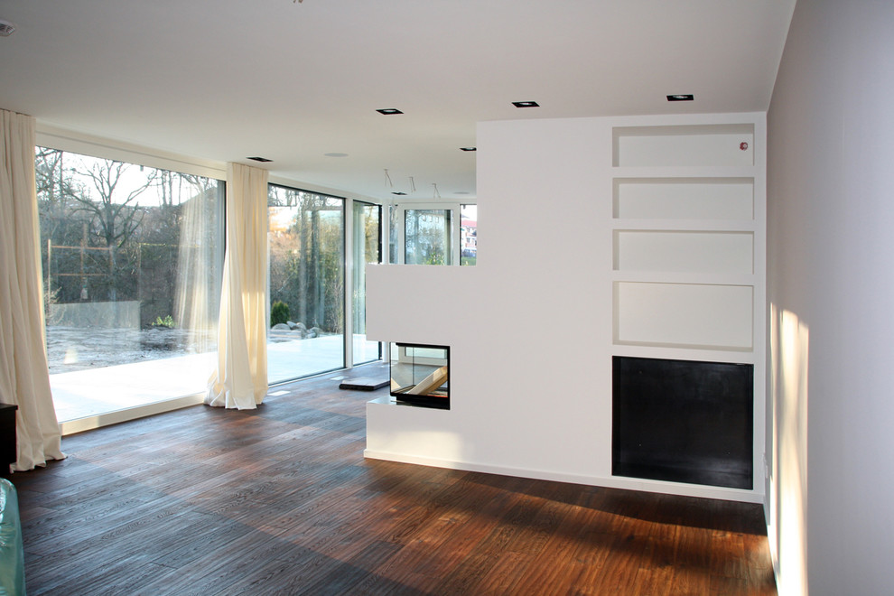 Пример оригинального дизайна: большая открытая гостиная комната в современном стиле с белыми стенами, темным паркетным полом, печью-буржуйкой, фасадом камина из штукатурки, телевизором на стене и коричневым полом