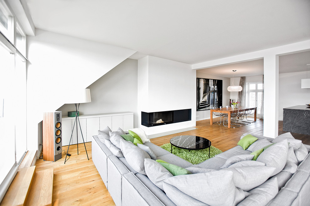 Imagen de sala de estar abierta contemporánea grande sin televisor con paredes blancas, suelo de madera en tonos medios, todas las chimeneas y marco de chimenea de yeso