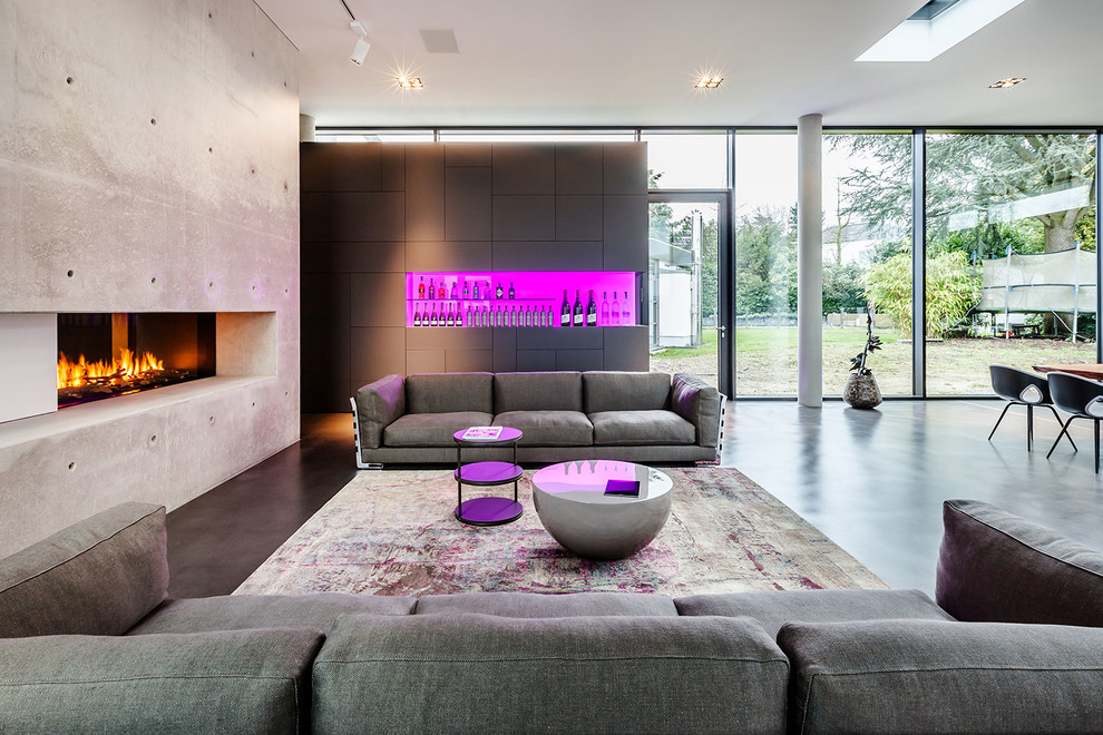 Foto de sala de estar con barra de bar actual extra grande con paredes grises, suelo de cemento, chimenea de doble cara y marco de chimenea de piedra