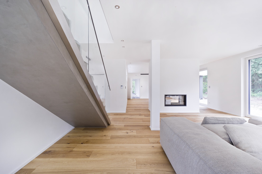 Cette image montre un grand salon minimaliste ouvert avec un mur blanc, un sol en bois brun, une cheminée double-face, un manteau de cheminée en plâtre, un téléviseur fixé au mur et un sol marron.