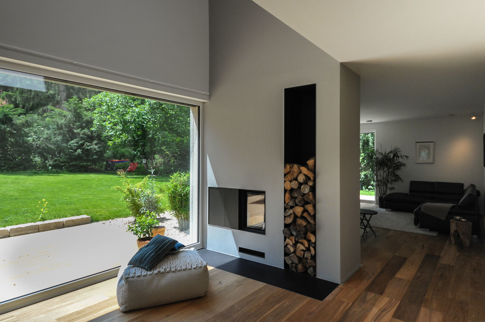 Modelo de salón abierto contemporáneo de tamaño medio con suelo de madera en tonos medios, chimenea de doble cara, marco de chimenea de yeso, paredes blancas y suelo marrón
