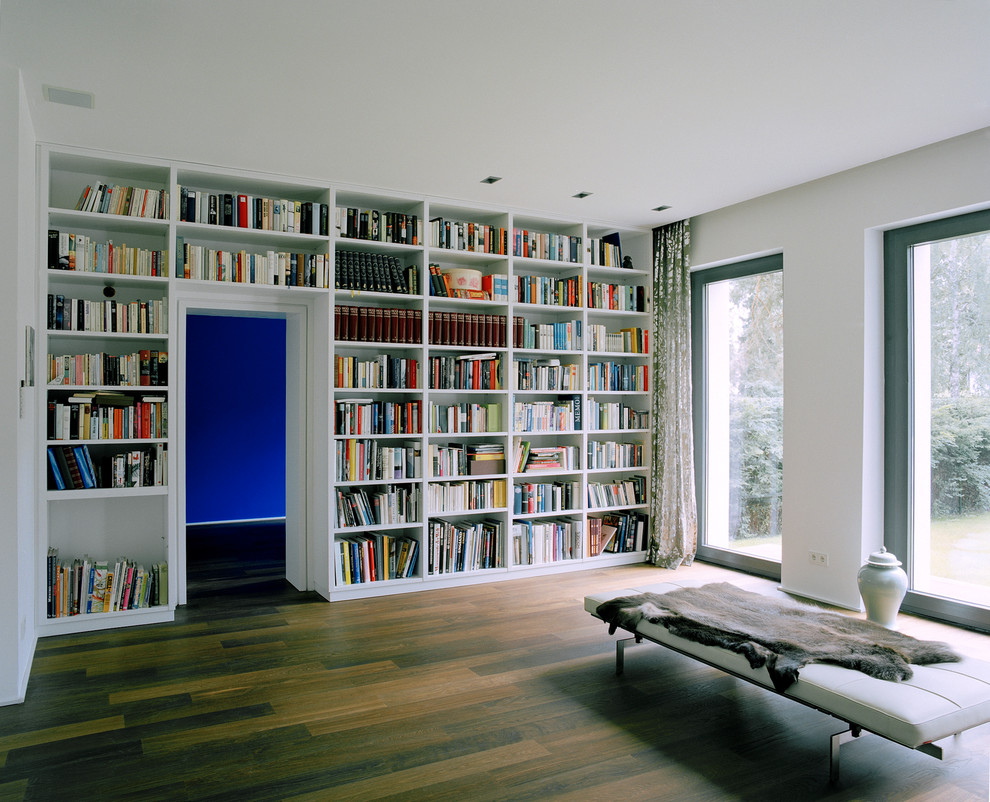 Modelo de biblioteca en casa abierta moderna grande con paredes blancas y suelo de madera en tonos medios