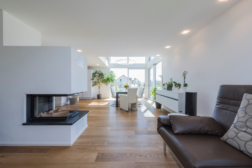 Modelo de salón abierto minimalista grande con paredes blancas, chimenea de doble cara, suelo de madera clara y marco de chimenea de baldosas y/o azulejos