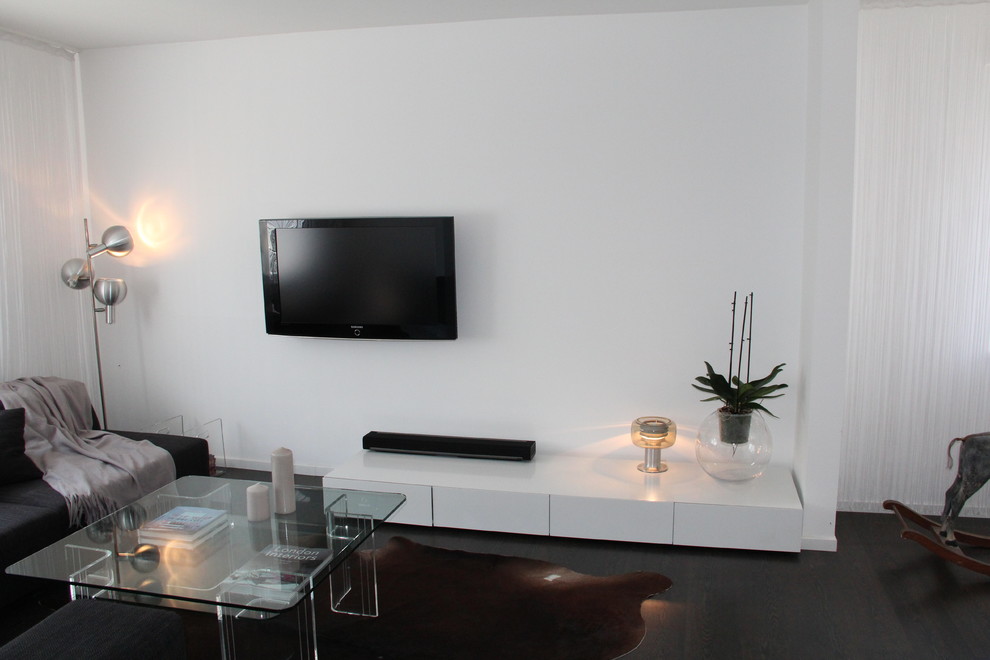 Modelo de sala de estar abierta contemporánea de tamaño medio sin chimenea con paredes blancas, suelo de madera oscura y televisor colgado en la pared