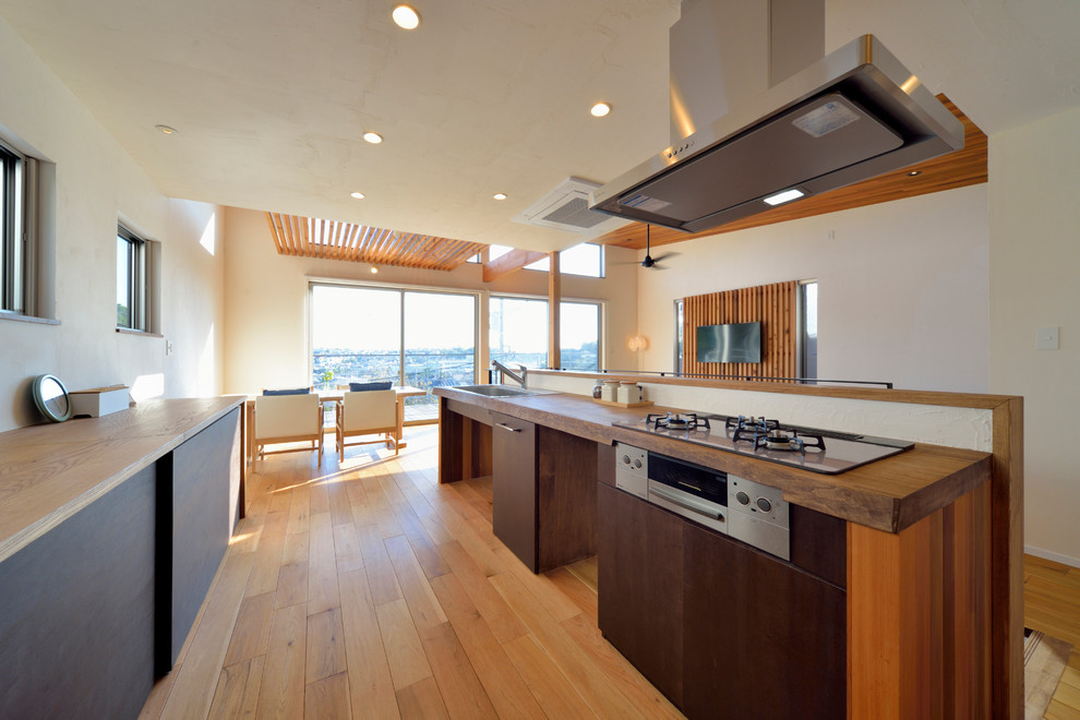Example of a trendy single-wall medium tone wood floor and brown floor open concept kitchen design in Other with a single-bowl sink, flat-panel cabinets, dark wood cabinets, wood countertops, white backsplash and an island