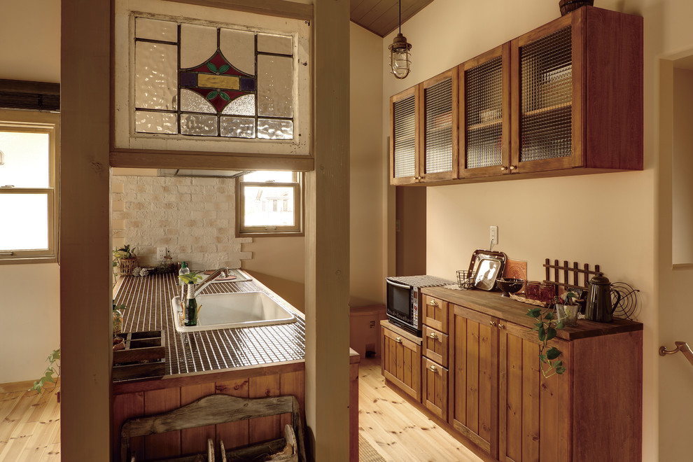 На фото: параллельная кухня-гостиная в стиле кантри с накладной мойкой, фасадами с утопленной филенкой, фасадами цвета дерева среднего тона, столешницей из плитки, светлым паркетным полом, островом и коричневым полом с