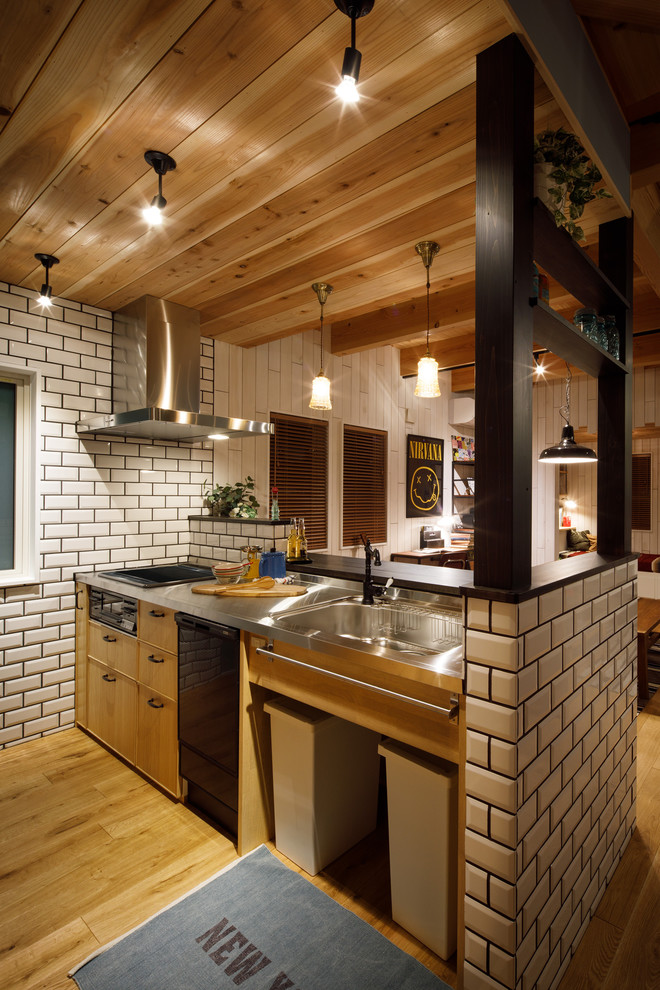 Exempel på ett mellanstort maritimt svart linjärt svart kök med öppen planlösning, med en undermonterad diskho, luckor med profilerade fronter, skåp i ljust trä, bänkskiva i rostfritt stål, vitt stänkskydd, stänkskydd i porslinskakel, ljust trägolv, en halv köksö och brunt golv