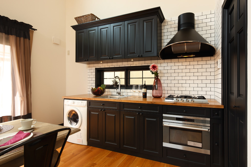 Exempel på ett 50 tals brun linjärt brunt kök med öppen planlösning, med en enkel diskho, luckor med upphöjd panel, skåp i mellenmörkt trä, träbänkskiva, vitt stänkskydd, stänkskydd i tunnelbanekakel, svarta vitvaror, mellanmörkt trägolv, en halv köksö och brunt golv