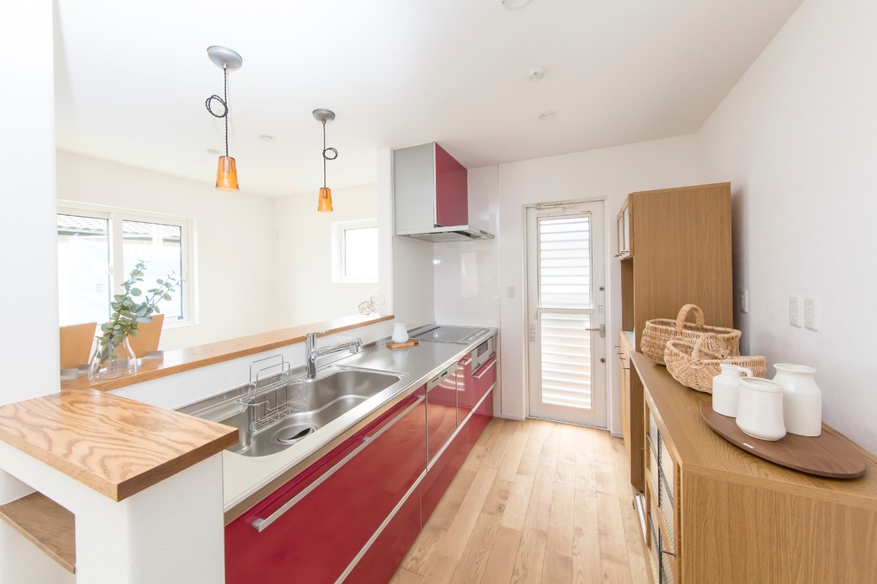 Contemporary galley open plan kitchen in Other with a single-bowl sink, flat-panel cabinets, red cabinets, stainless steel worktops, red splashback, light hardwood flooring and an island.