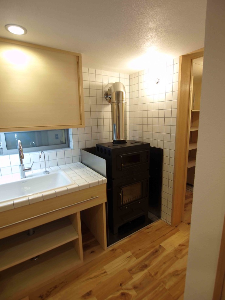 Klassische Wohnküche mit Einbauwaschbecken, flächenbündigen Schrankfronten, hellen Holzschränken, Arbeitsplatte aus Fliesen, Küchenrückwand in Weiß, Rückwand aus Glasfliesen, schwarzen Elektrogeräten, braunem Holzboden und Halbinsel in Tokio