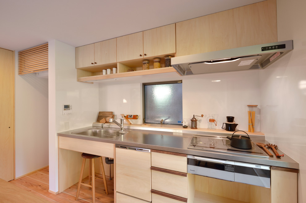 Photo of a world-inspired single-wall open plan kitchen in Other with an integrated sink, flat-panel cabinets, light wood cabinets, stainless steel worktops, white splashback, light hardwood flooring, a breakfast bar, brown floors and beige worktops.