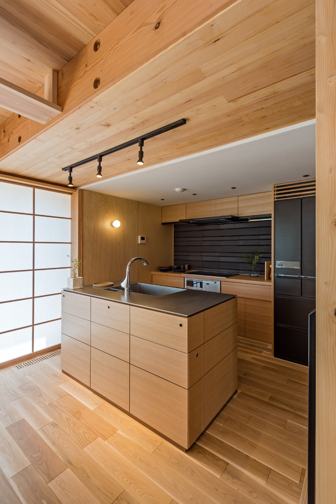 Modelo de cocina de estilo zen abierta con fregadero integrado, armarios con paneles lisos, puertas de armario de madera oscura, salpicadero marrón, electrodomésticos negros, suelo de madera en tonos medios y una isla