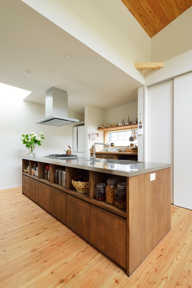 Idéer för att renovera ett litet funkis linjärt kök med öppen planlösning, med en integrerad diskho, bruna skåp, bänkskiva i rostfritt stål, rostfria vitvaror, mellanmörkt trägolv, beiget golv, släta luckor och en köksö