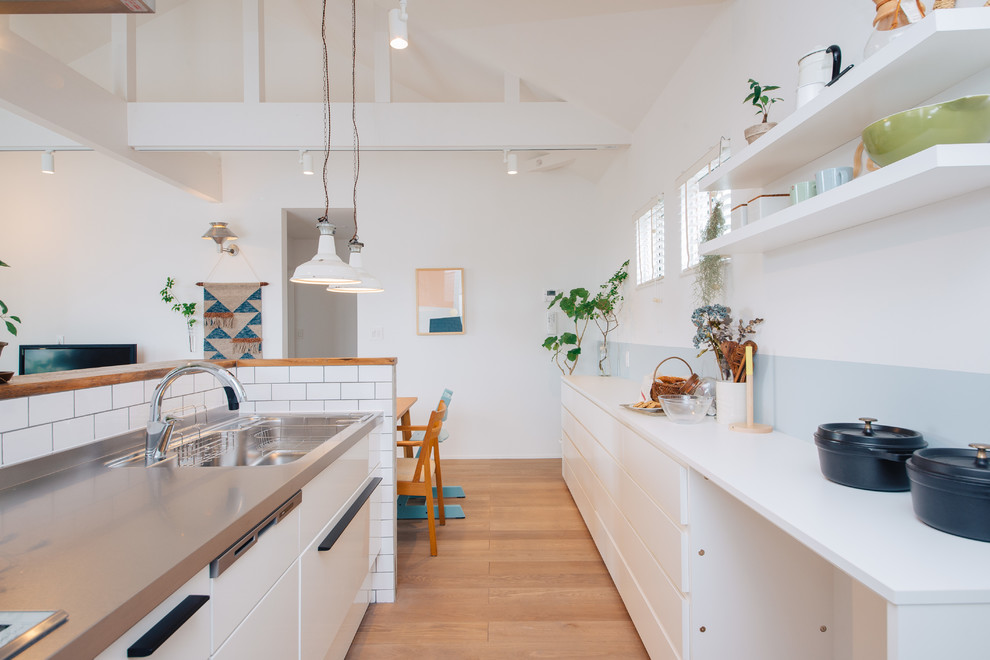 Exempel på ett skandinaviskt vit linjärt vitt kök med öppen planlösning, med en undermonterad diskho, vita skåp, bänkskiva i rostfritt stål, vitt stänkskydd, stänkskydd i porslinskakel, plywoodgolv och en köksö