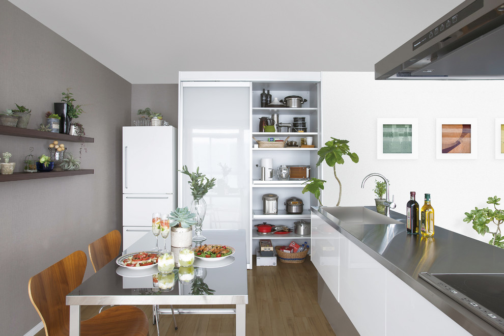 Inredning av ett modernt linjärt kök och matrum, med en integrerad diskho, släta luckor, vita skåp, bänkskiva i rostfritt stål, vita vitvaror, mellanmörkt trägolv och en halv köksö