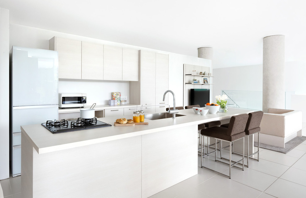 Пример оригинального дизайна: кухня-гостиная в стиле модернизм с плоскими фасадами, белым фартуком, островом, одинарной мойкой, светлыми деревянными фасадами и белой техникой