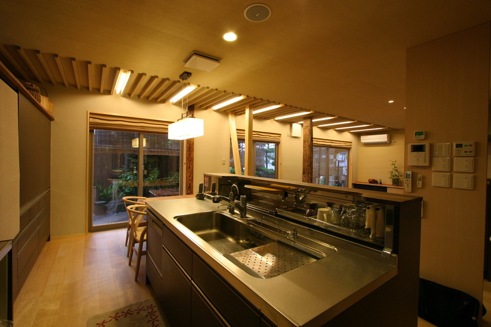 Asiatisk inredning av ett mellanstort grå grått kök med öppen planlösning, med en enkel diskho, släta luckor, bruna skåp, bänkskiva i rostfritt stål, vitt stänkskydd, svarta vitvaror, mellanmörkt trägolv, en köksö och beiget golv