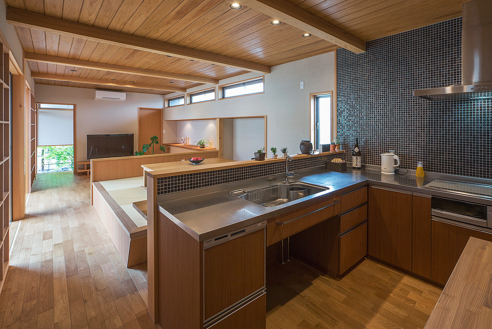 Asiatisk inredning av ett kök, med en integrerad diskho, släta luckor, skåp i mellenmörkt trä, bänkskiva i rostfritt stål, svart stänkskydd, stänkskydd i mosaik, rostfria vitvaror och mellanmörkt trägolv