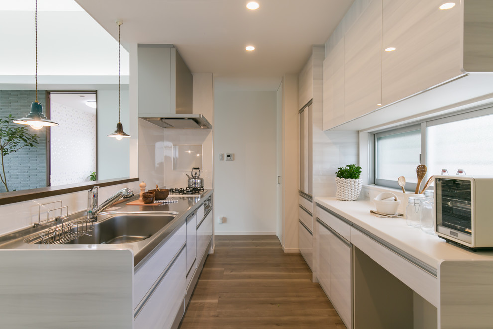 Foto på ett funkis kök, med en enkel diskho, släta luckor, grå skåp, bänkskiva i rostfritt stål, vitt stänkskydd, mellanmörkt trägolv, en köksö och brunt golv