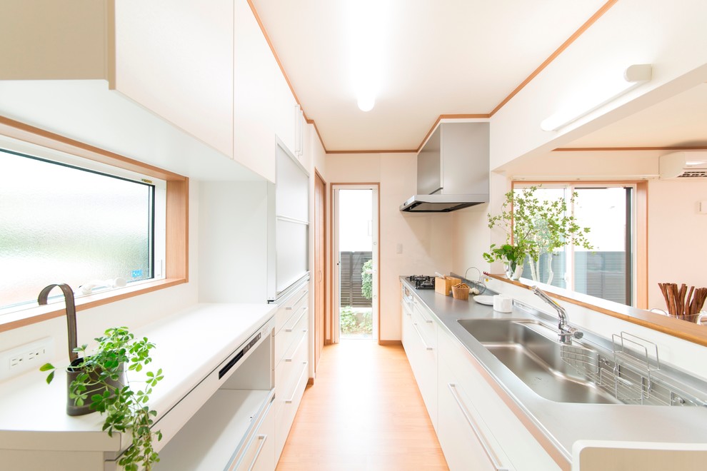 Foto på ett funkis linjärt kök, med en integrerad diskho, släta luckor, vita skåp, bänkskiva i rostfritt stål, ljust trägolv och vitt stänkskydd