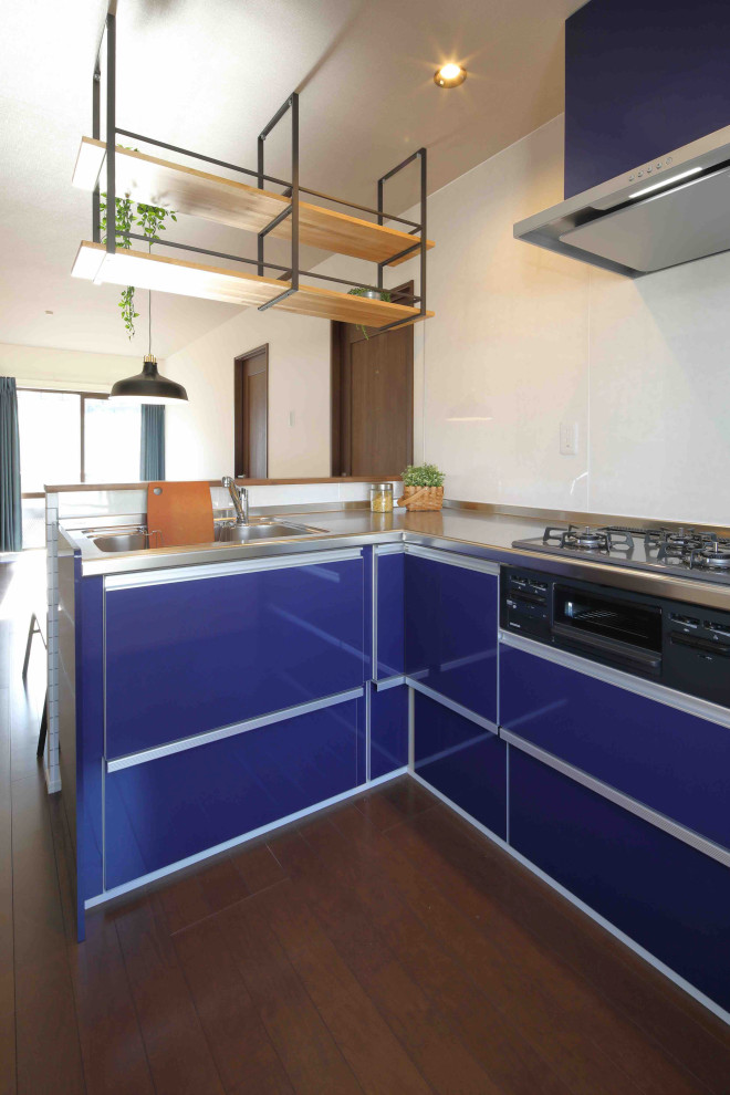 На фото: угловая кухня-гостиная в стиле ретро с монолитной мойкой, плоскими фасадами, синими фасадами, столешницей из нержавеющей стали, белым фартуком, полом из фанеры и коричневым полом